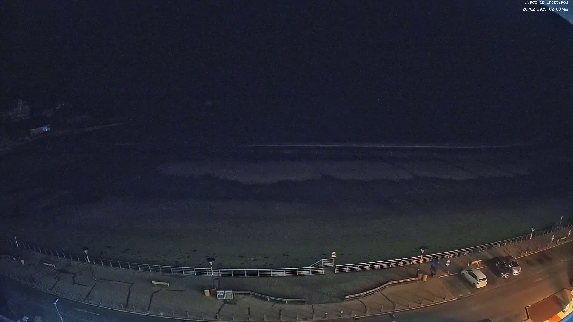 A nighttime view of a dark beach with a calm ocean and a few cars parked along a boardwalk.