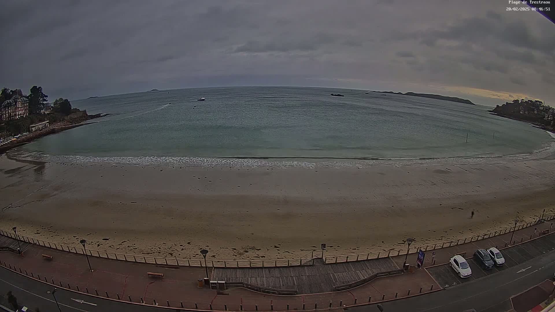 A sandy beach with calm waves under an overcast sky is seen from an elevated viewpoint, showing a boardwalk and parked cars along the shoreline.