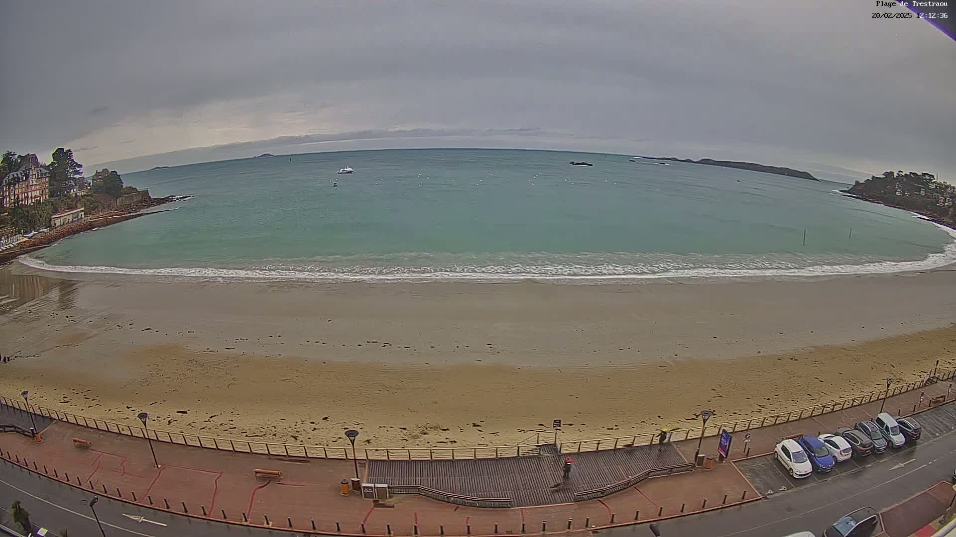 A sandy beach with calm, teal-green water under an overcast sky is viewed from above, showing a boardwalk and parked cars along the shore.
