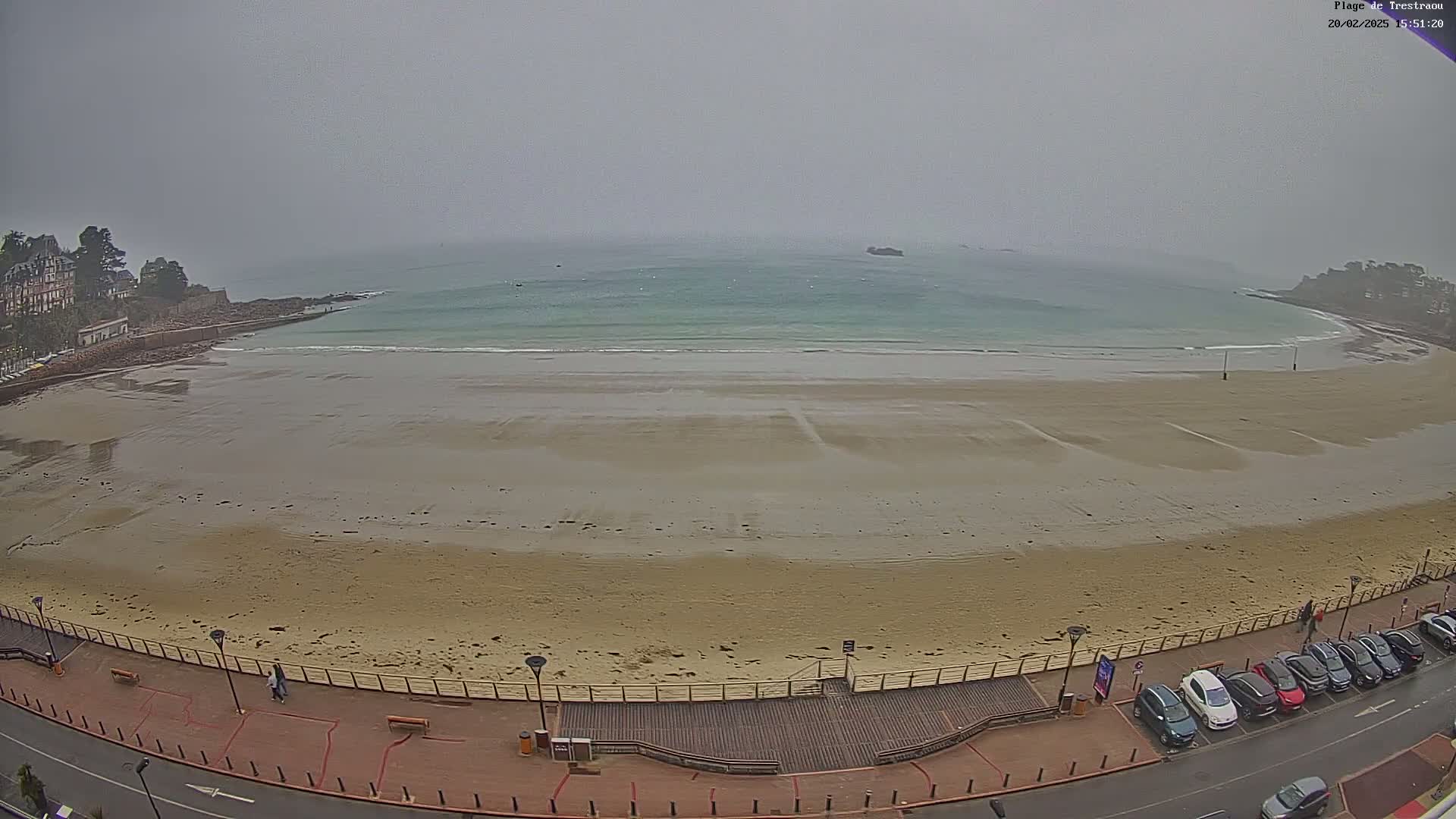 A wide sandy beach stretches out to a calm, light-green sea under an overcast sky.