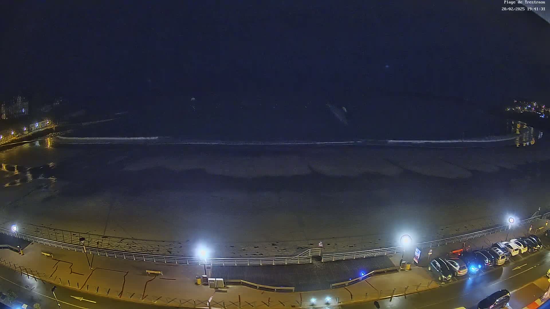 A nighttime, high-angle view of a mostly empty beach with small waves lapping the shore, illuminated by streetlights along a boardwalk with parked cars.