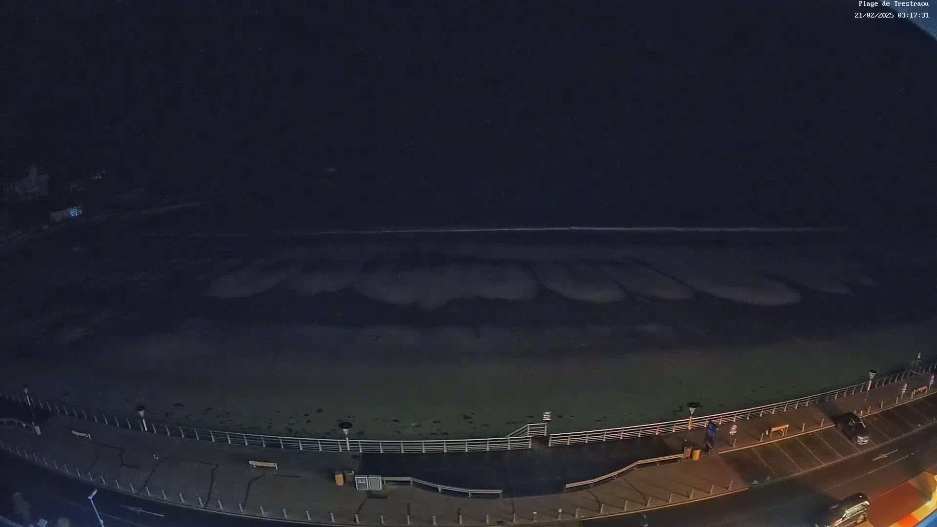 A dark, nighttime view shows a mostly empty beach with a boardwalk and a few cars parked nearby.