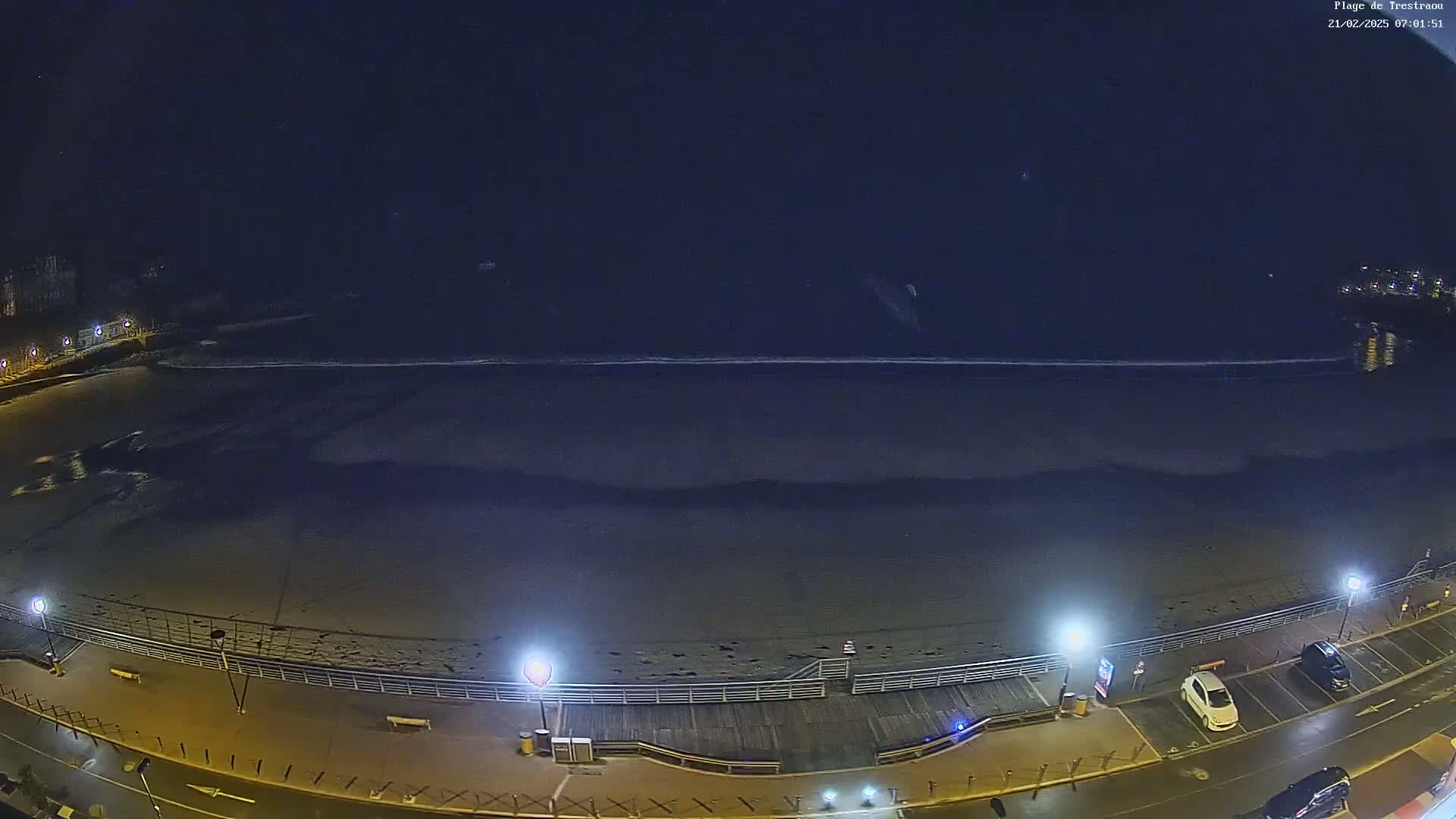 A dark, nighttime beach scene shows a mostly empty shore with calm waves under a clear, starless sky, illuminated by streetlights along a boardwalk and parking area.