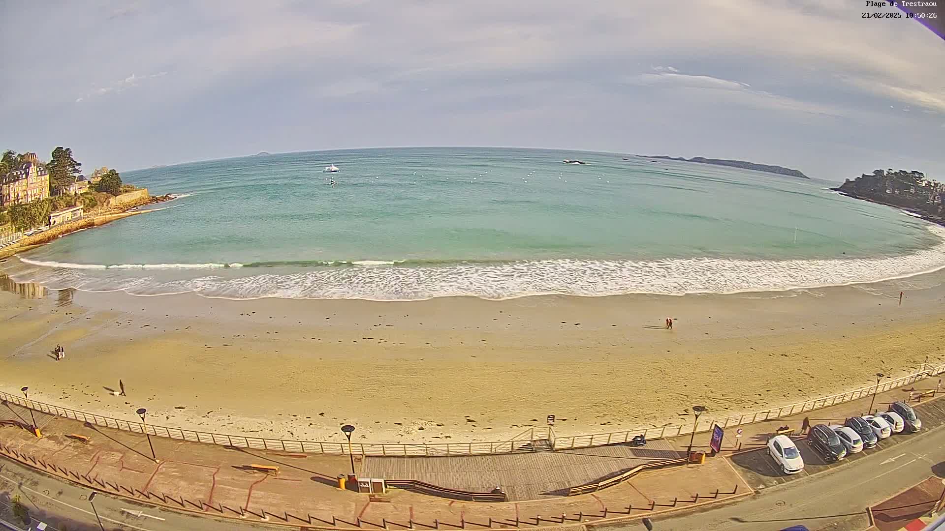 A sandy beach with gentle waves under a partly cloudy sky is bordered by a paved walkway and a row of parked cars.