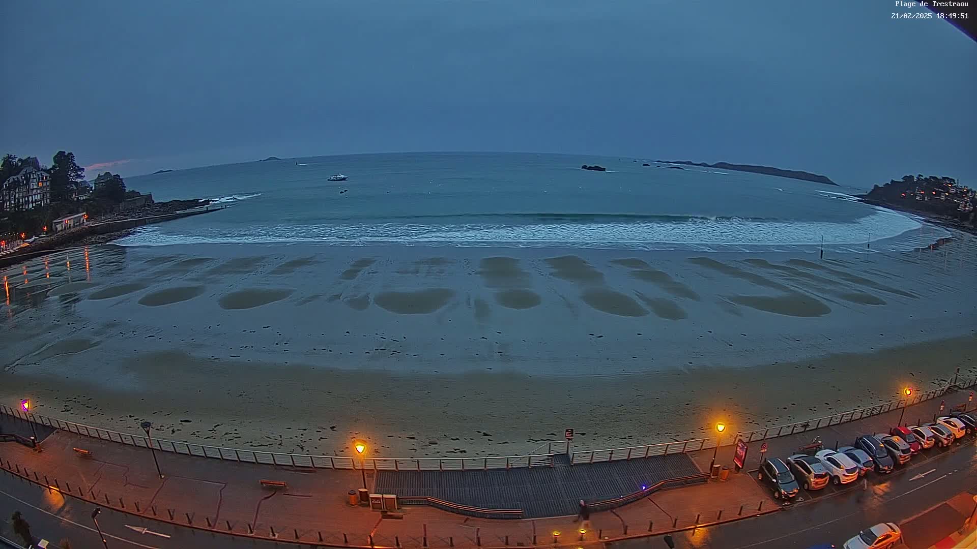 A sandy beach at low tide under a dusky sky, with gentle waves lapping the shore and a row of cars parked along a boardwalk at the edge of the beach.