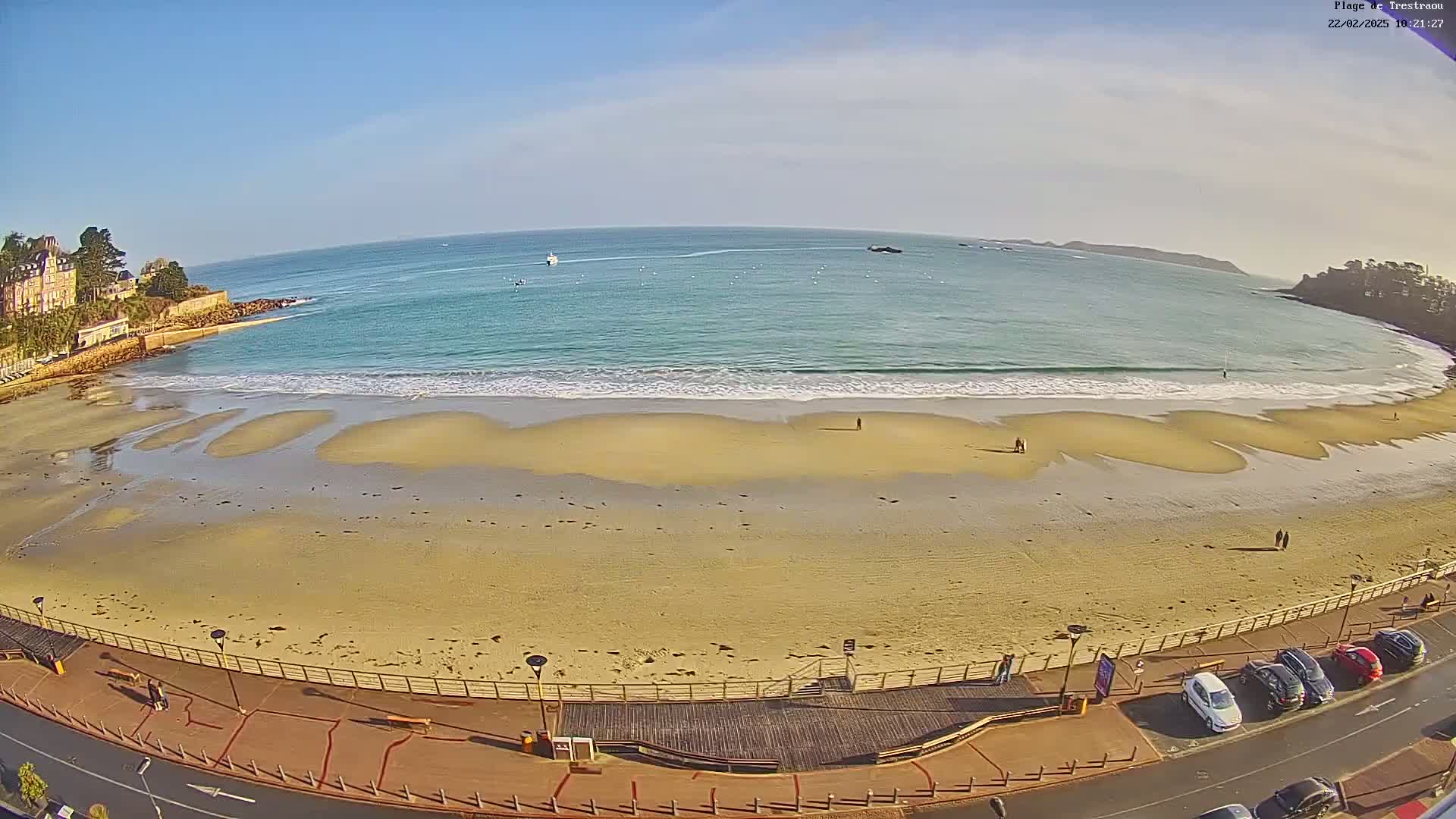 A sandy beach with gentle waves under a clear blue sky is bordered by a paved walkway and a row of parked cars.