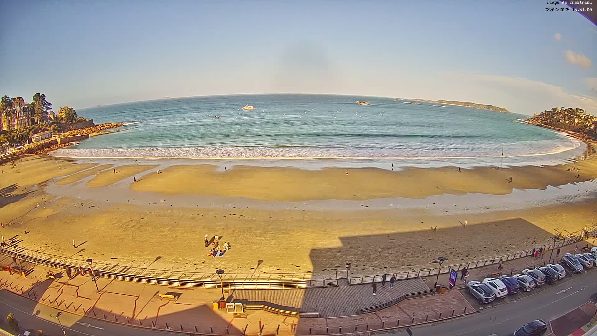 A sandy beach with gentle waves under a clear blue sky, overlooked by a boardwalk and a row of parked cars.