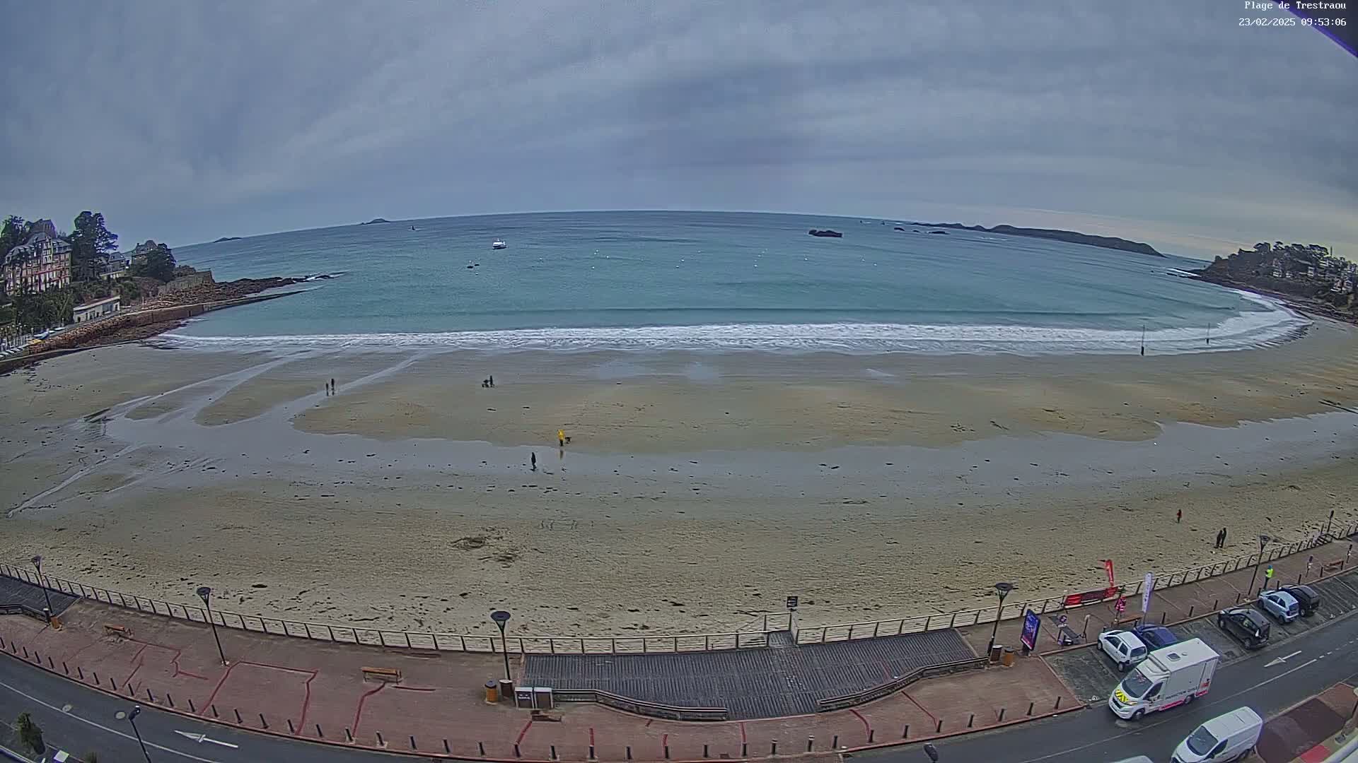 An overcast day shows a sandy beach with a few people walking along the shore, small waves lapping at the sand, and a calm ocean extending to distant islands.