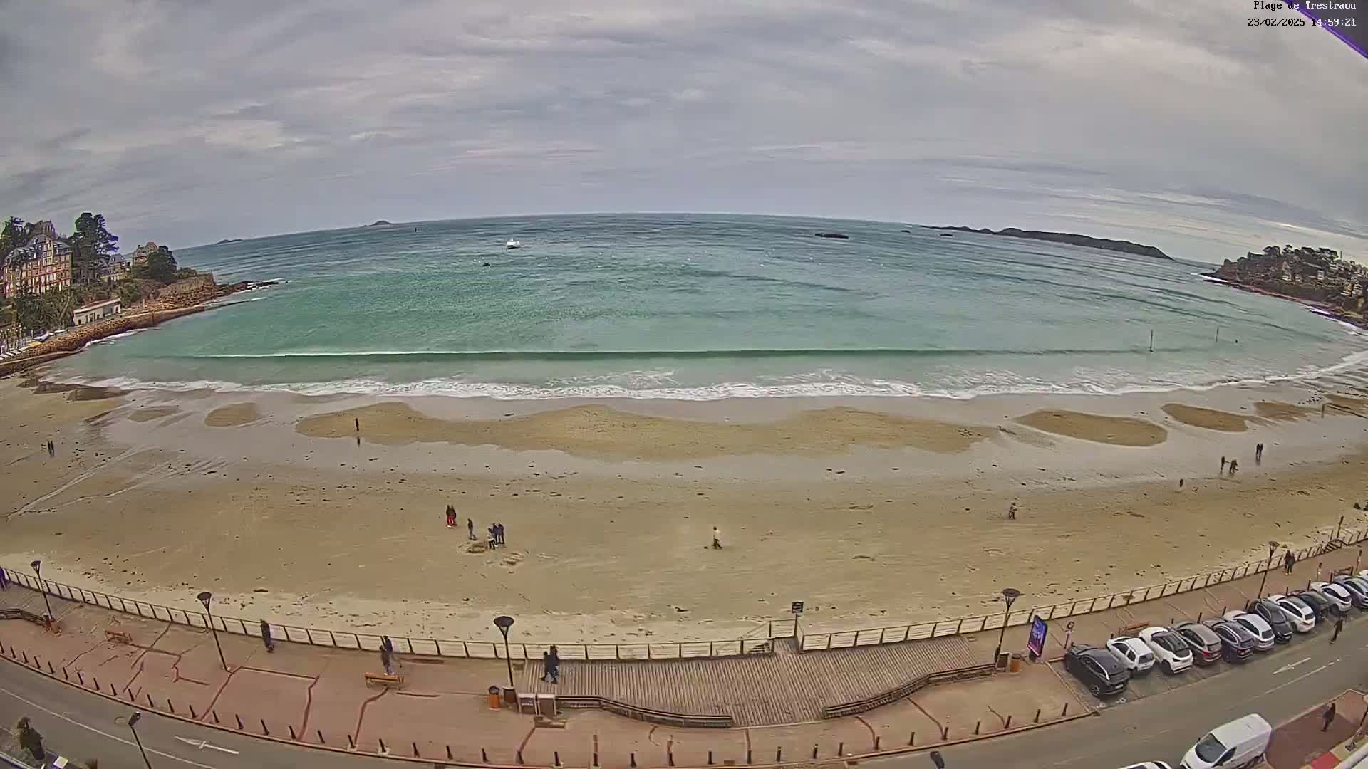 An overcast day shows a sandy beach with small waves lapping the shore, a few people walking on the sand, and cars parked along a roadway next to a boardwalk.