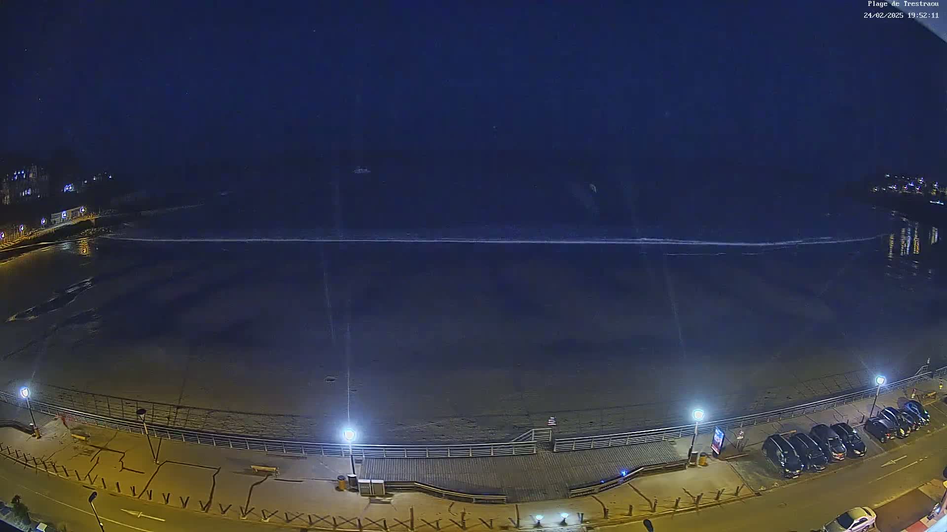 A dark beach at night with a calm ocean and several cars parked along a boardwalk.