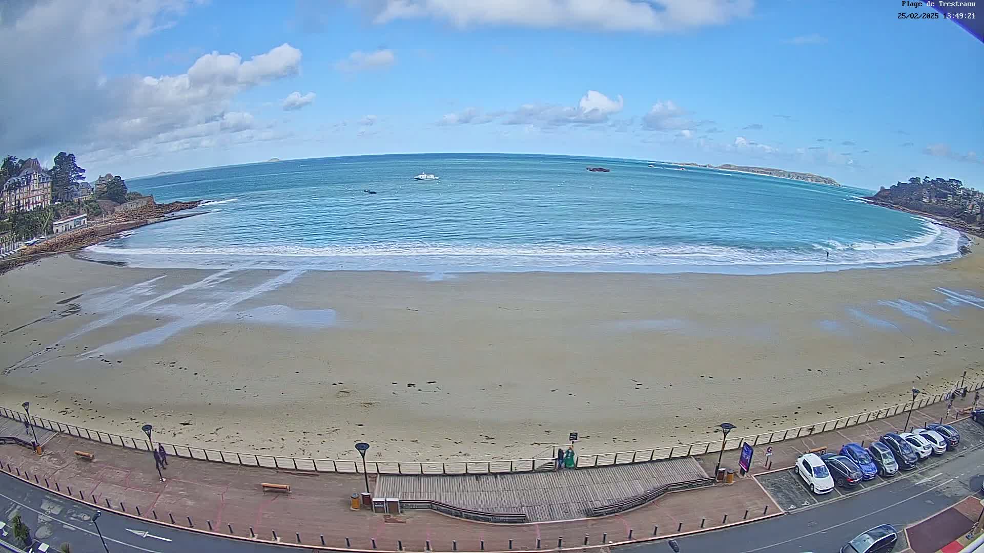 A sandy beach with small waves laps the shore under a partly cloudy blue sky; several cars are parked along a road next to the beach, and a few boats are visible in the distance on the water.