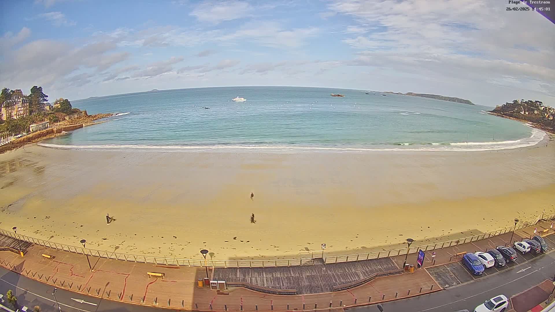 A mostly sunny day shows a long sandy beach with a few people walking, a boardwalk, and a parking lot with cars, overlooking a calm ocean with a few boats.