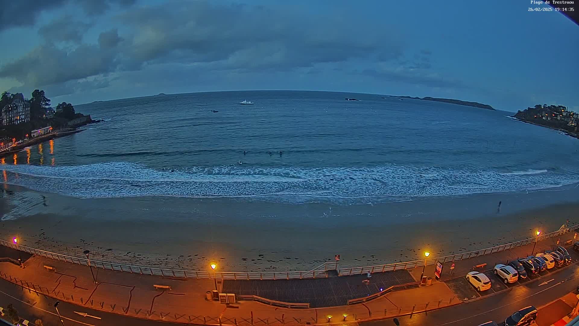 A mostly cloudy twilight scene shows a sandy beach with gentle waves lapping the shore, a boardwalk with parked cars, and buildings in the background.