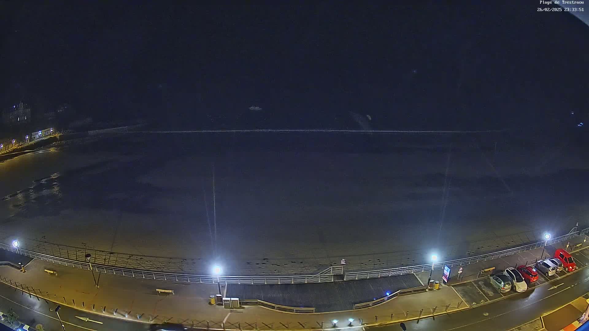 A dark, nighttime view of a beach with a boardwalk, streetlights, and a few parked cars.
