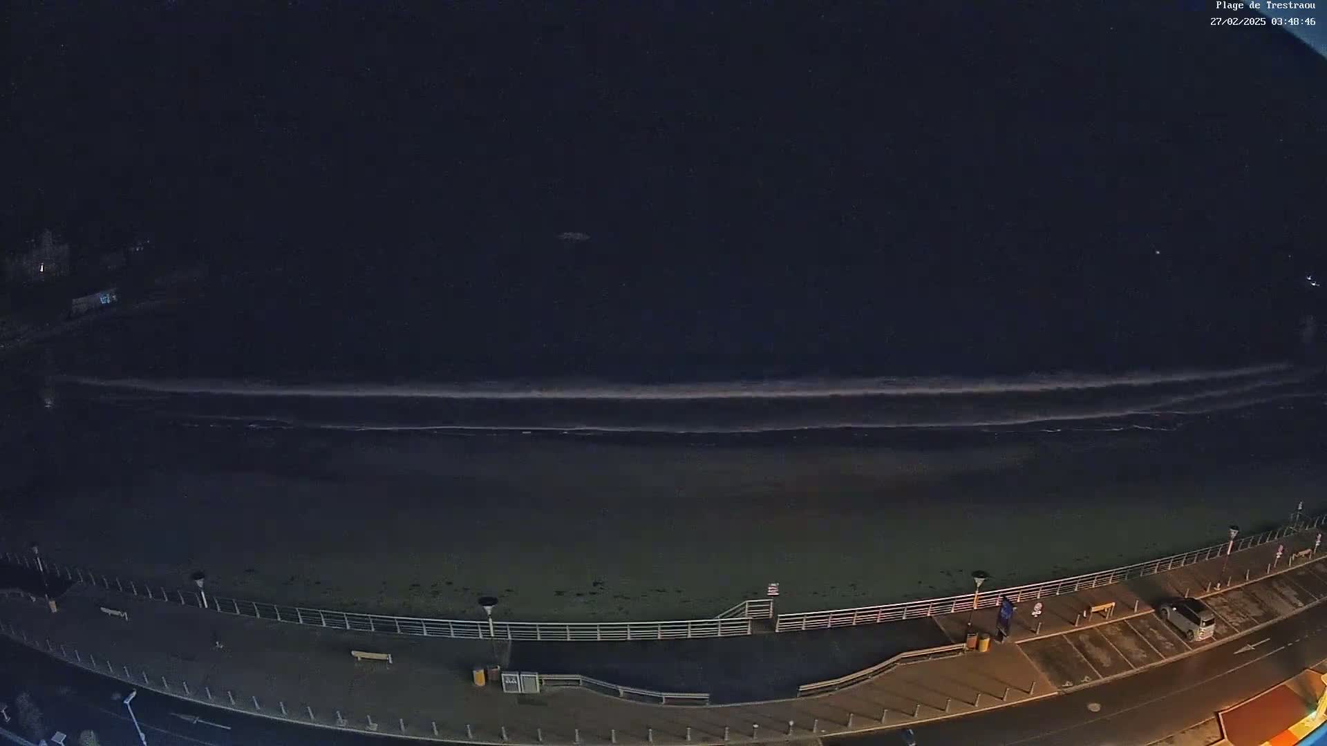 A dark, nighttime beach scene shows gentle waves lapping the shore, a paved walkway, and a few cars parked nearby.