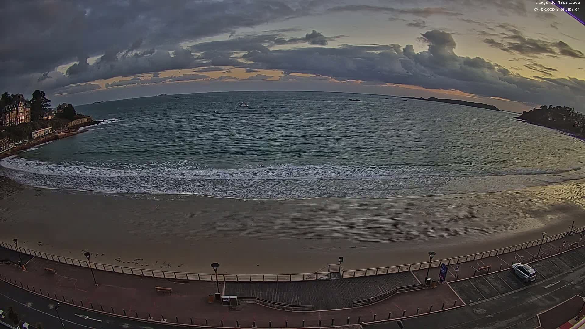A sandy beach with gentle waves laps the shore under a partly cloudy sky at sunrise or sunset, with a boardwalk and buildings visible in the background.