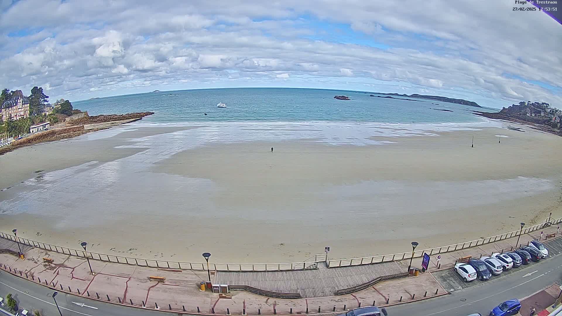 A mostly cloudy day reveals a wide sandy beach with a boardwalk, a few cars parked nearby, and calm ocean waters stretching to the horizon.