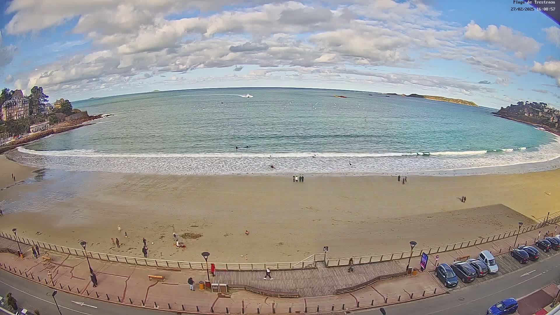 A sandy beach with gentle waves laps the shore under a partly cloudy sky, with a few people scattered on the sand and boardwalk, and cars parked along a nearby road.