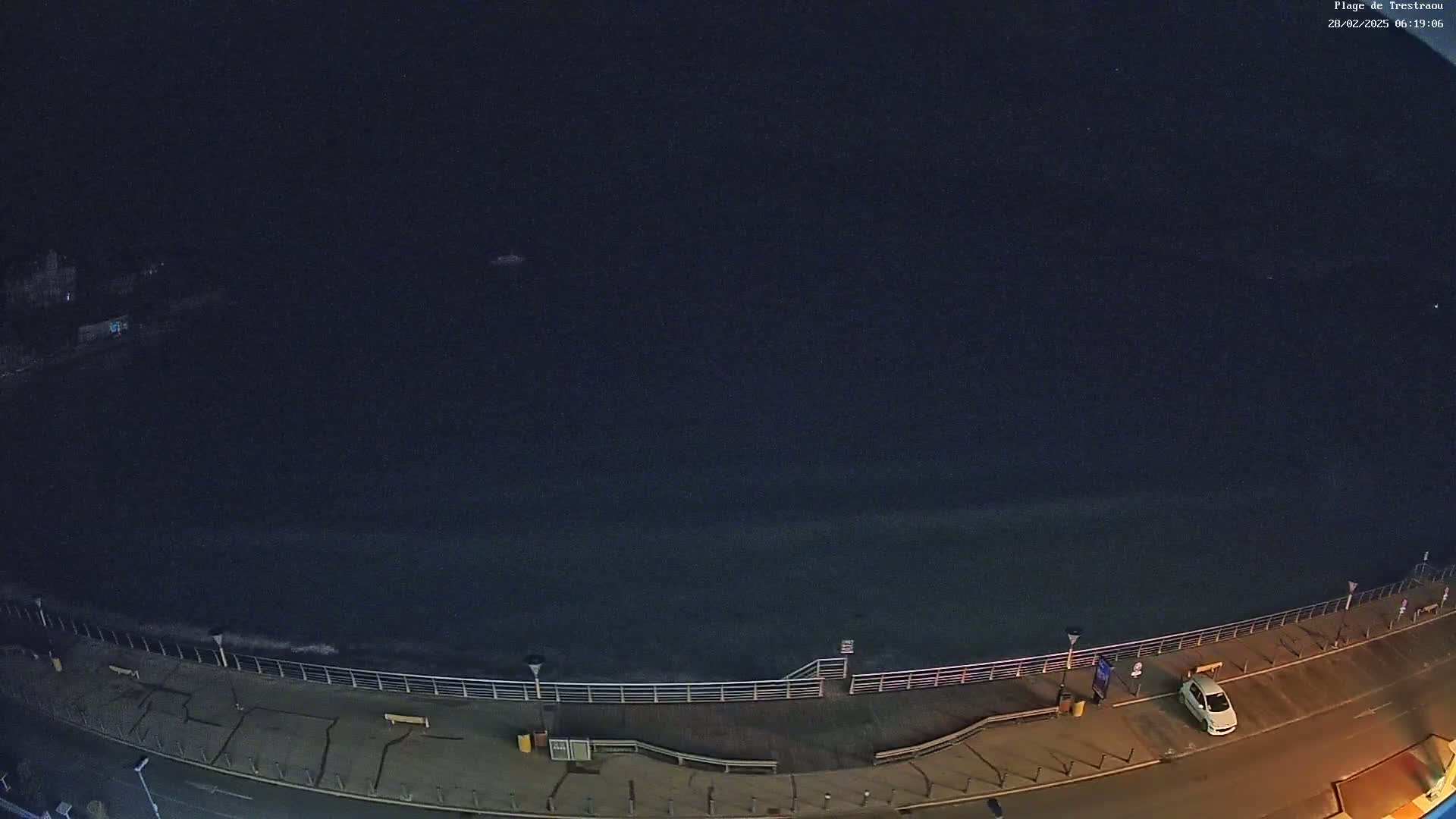 A nighttime, high-angle view shows a dark ocean meeting a paved walkway with a few parked cars and some people visible under dimly lit streetlights.