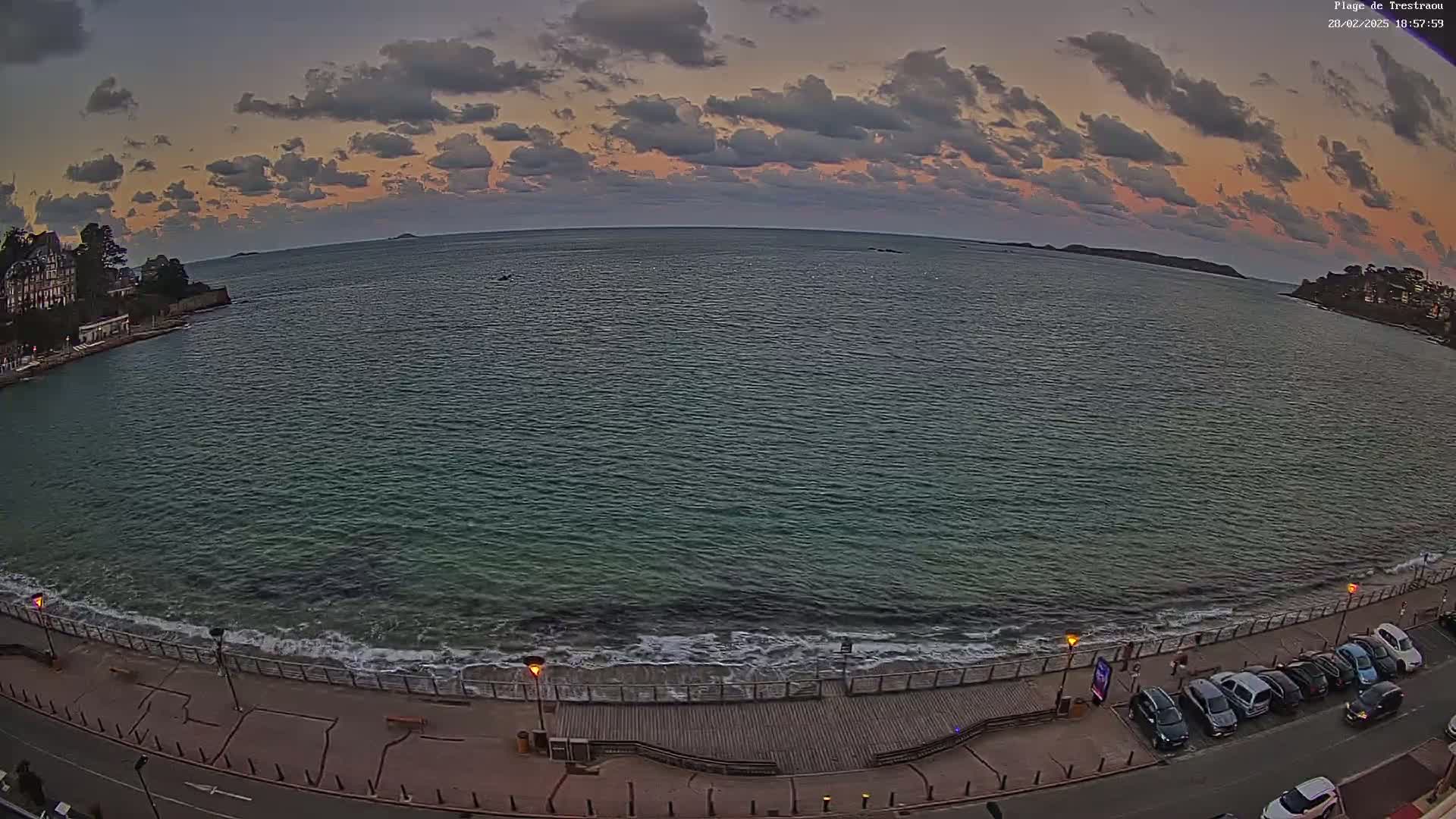 A wide shot of a calm sea at sunset with small waves lapping a boardwalk and a parking lot filled with cars.