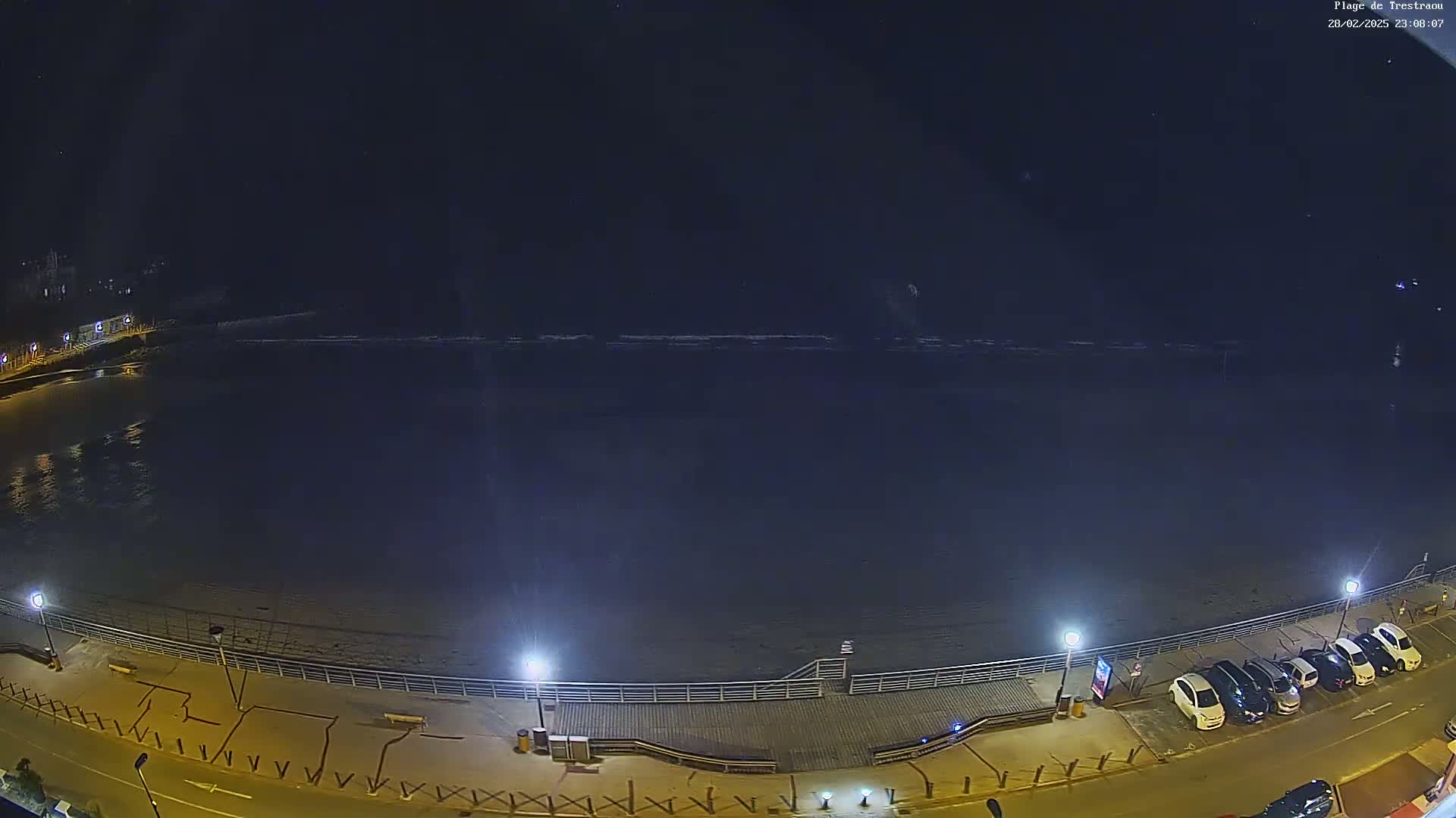 A nighttime, high-angle view shows a dark, calm ocean lapping a sandy beach with a boardwalk and parked cars along a street.