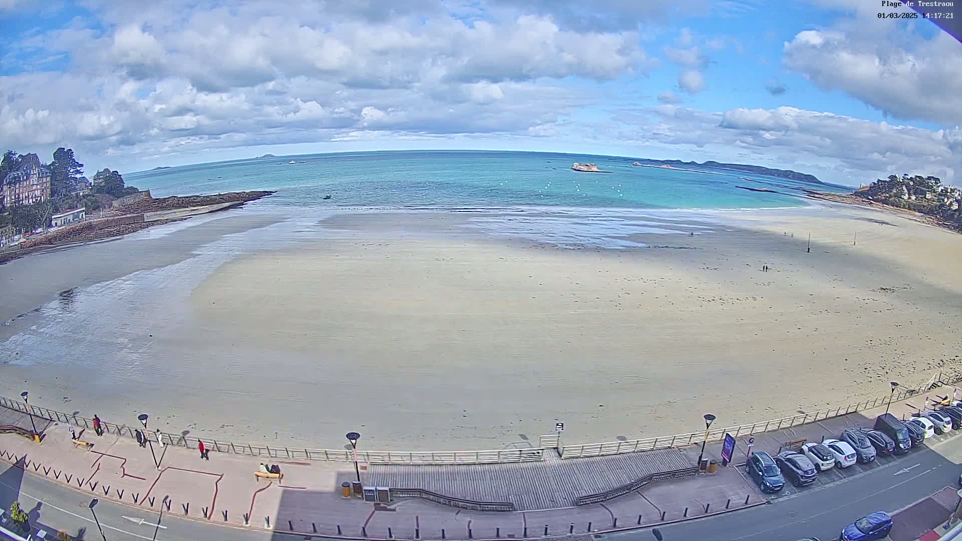 A mostly sandy beach stretches along a calm, turquoise ocean under a partly cloudy sky; a boardwalk and parked cars line the beach's edge.