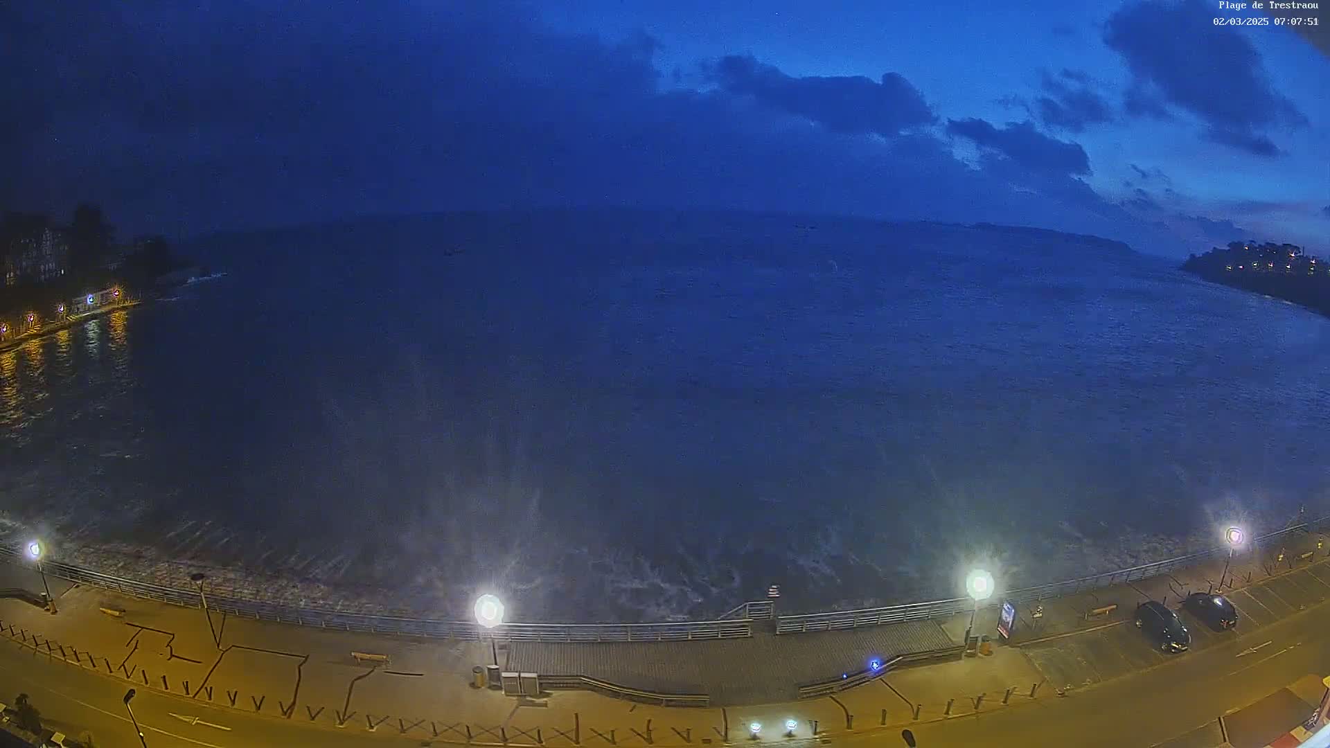 Under a dark, cloudy sky, waves crash against a shoreline at night, where a paved walkway and parking lot are illuminated by streetlights.