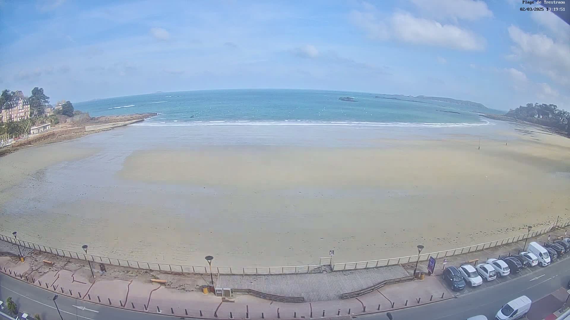 A mostly sunny day at a wide sandy beach with calm ocean waves, buildings, and parked cars visible along the shoreline.