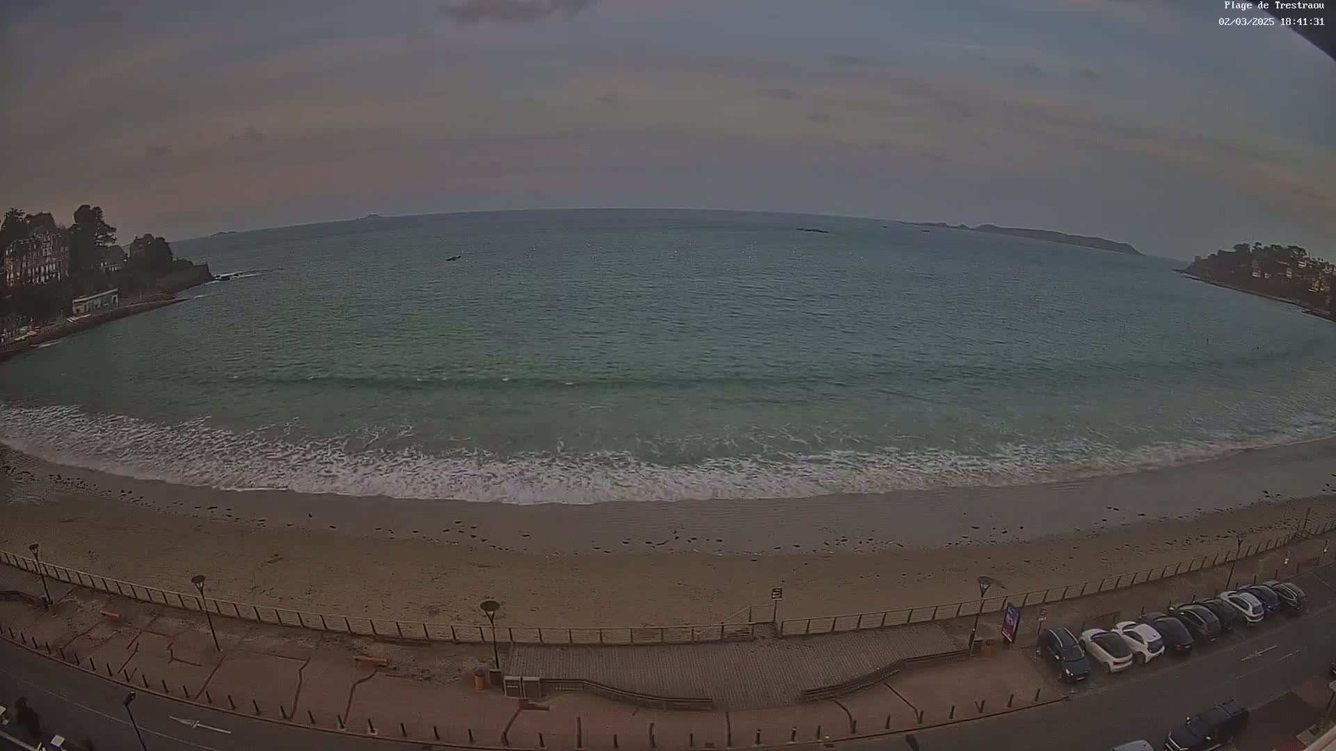 A sandy beach with gentle waves laps the shore under an overcast sky, with cars parked along a paved road overlooking the ocean.