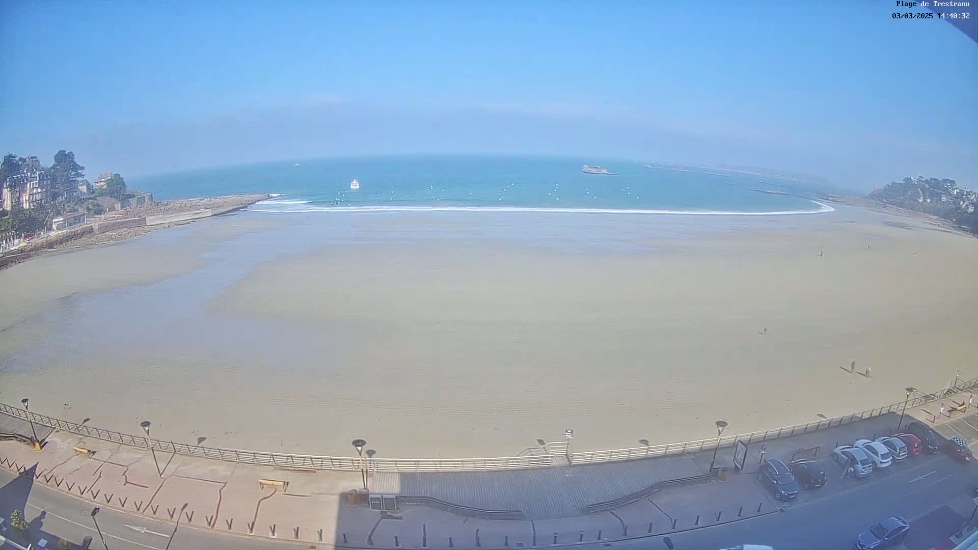 A mostly empty, sandy beach stretches out to a calm ocean under a clear blue sky, with a few cars parked along a boardwalk in the foreground.