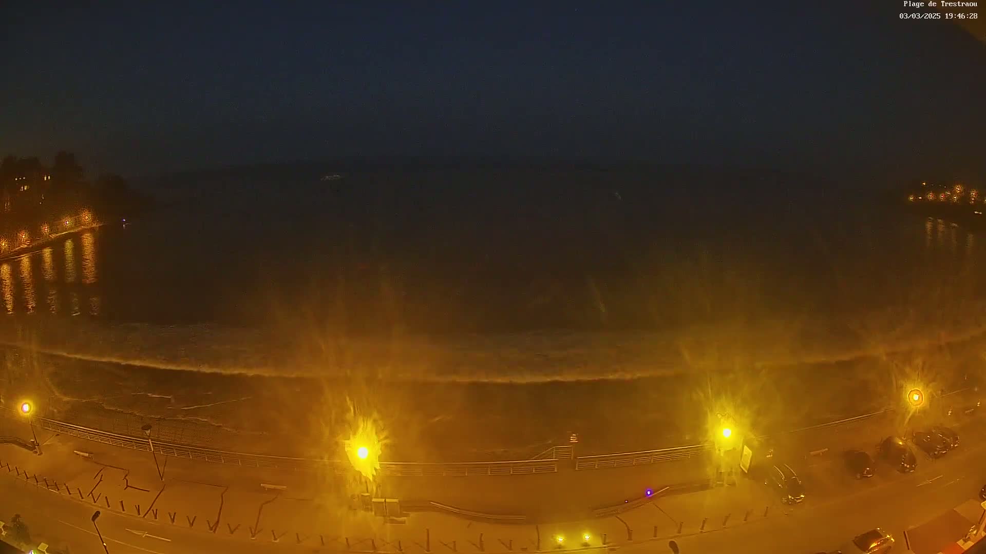 At night, large waves crash against a seawall illuminated by streetlights along a parked cars-lined roadway.