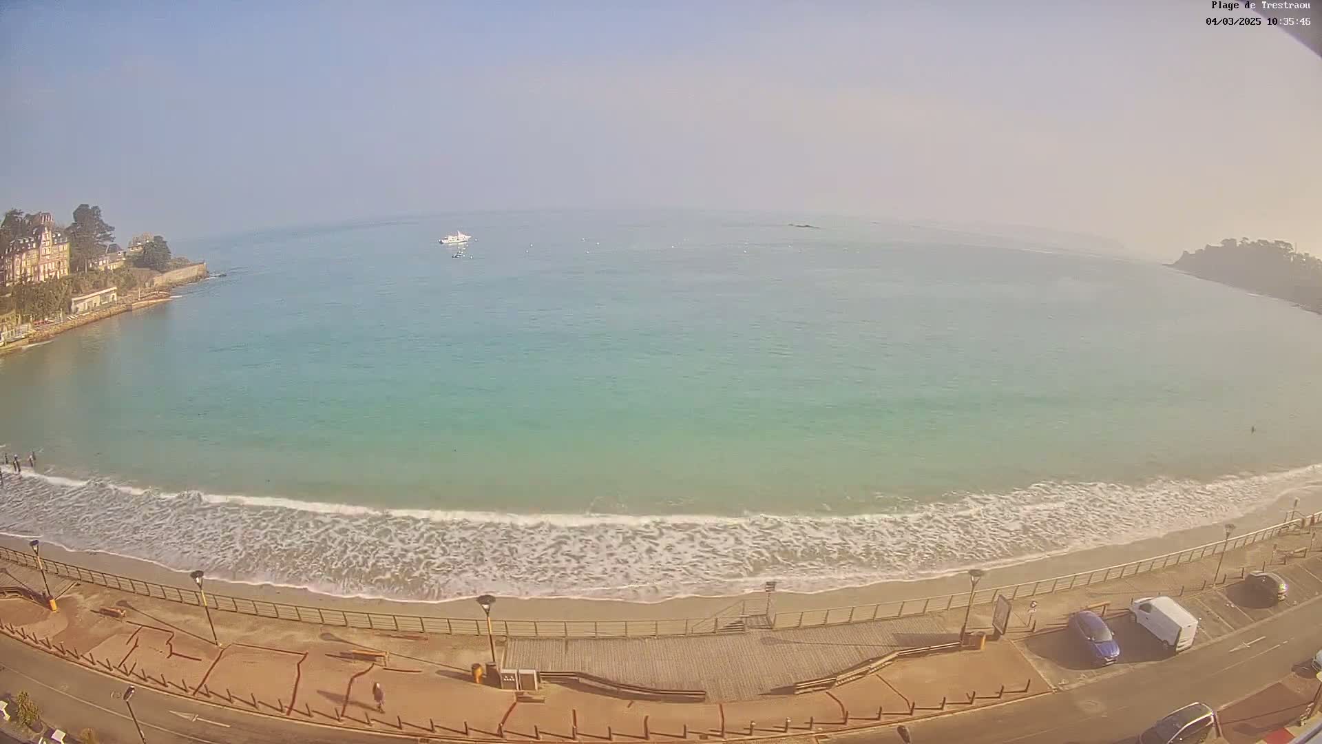A sandy beach with gentle waves laps the shore under a hazy sky, overlooked by a building and parking lot.