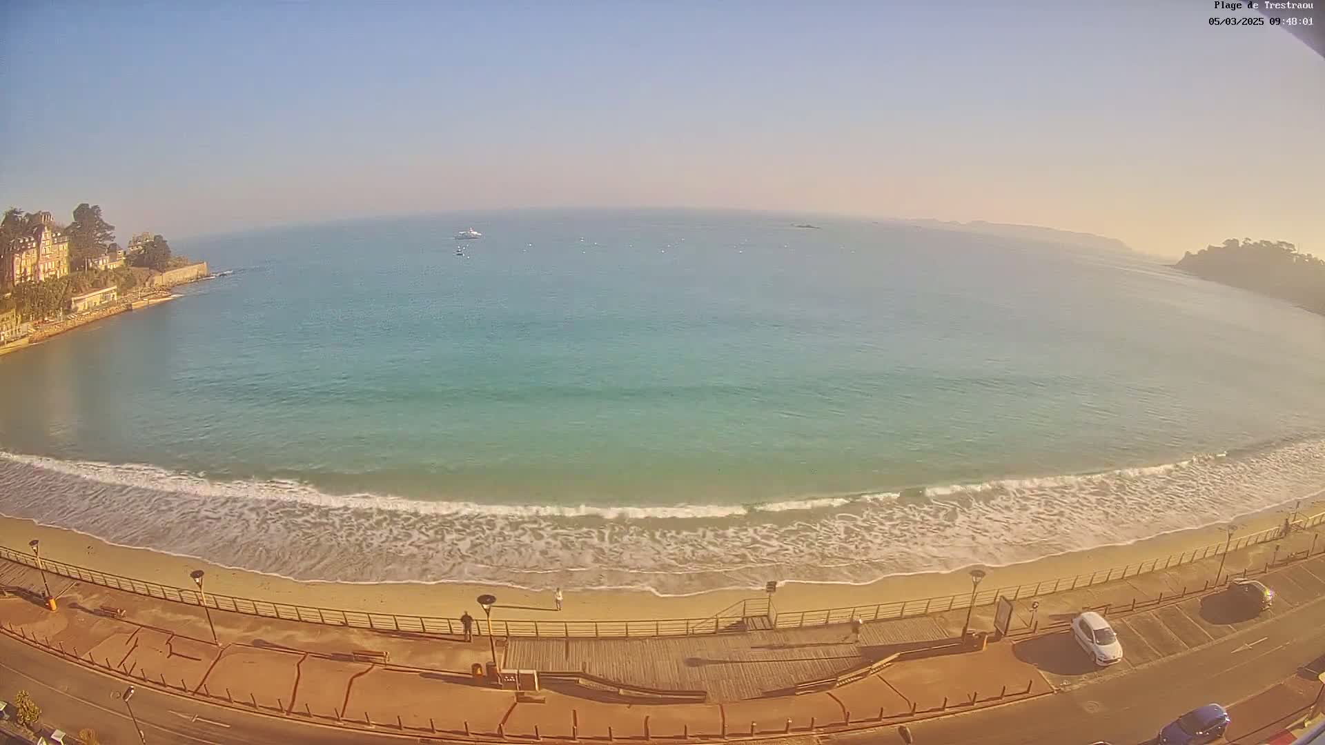 An aerial view shows a sandy beach with gentle waves lapping the shore under a clear, sunny sky, bordered by a boardwalk, a road, and buildings.