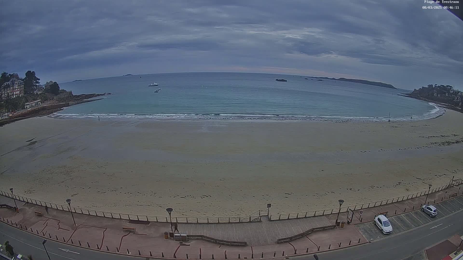 A sandy beach stretches along a calm, light-blue ocean under an overcast sky, with a boardwalk and parked cars visible along the shoreline.