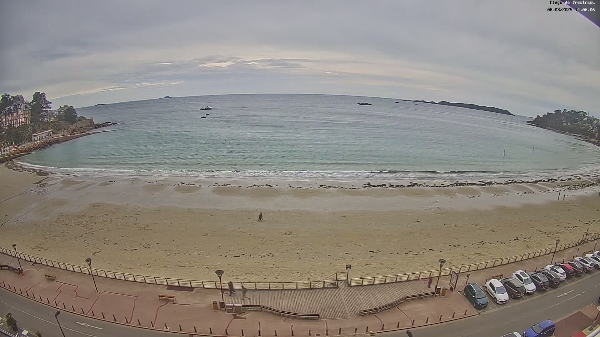 A wide shot of a sandy beach with calm, light-blue water under an overcast sky, showing a few people walking on the beach and cars parked along a road nearby.
