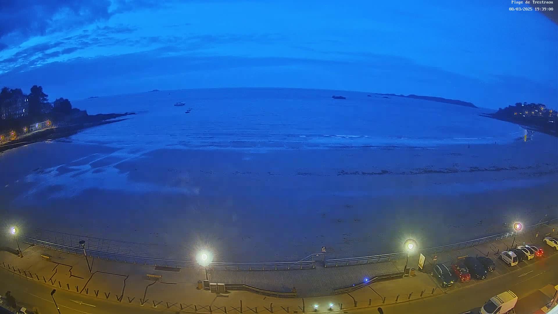 A wide, twilight-lit shot shows a beach at low tide, with a few cars parked along the road above and a few boats visible in the distance under a clear, dark blue sky.