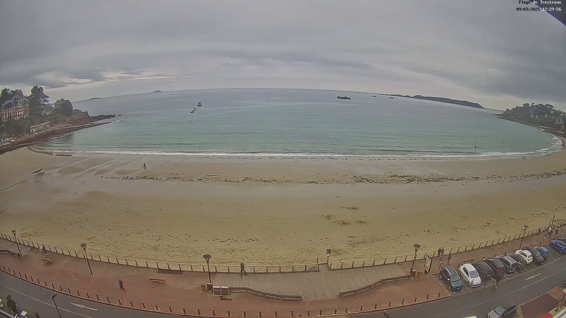 A sandy beach with calm, light-blue water under an overcast sky is viewed from above, showing a boardwalk, parked cars, and buildings in the background.