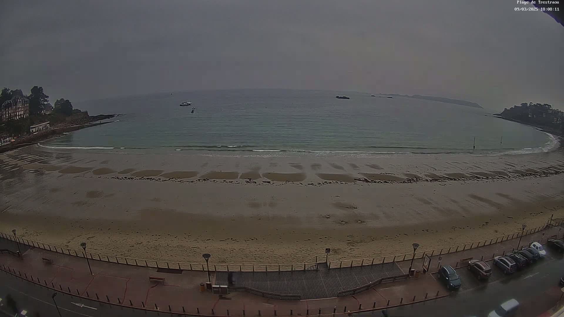 A sandy beach curves around a calm, gray-toned ocean under an overcast sky, with a few boats visible in the distance and cars parked along a boardwalk.