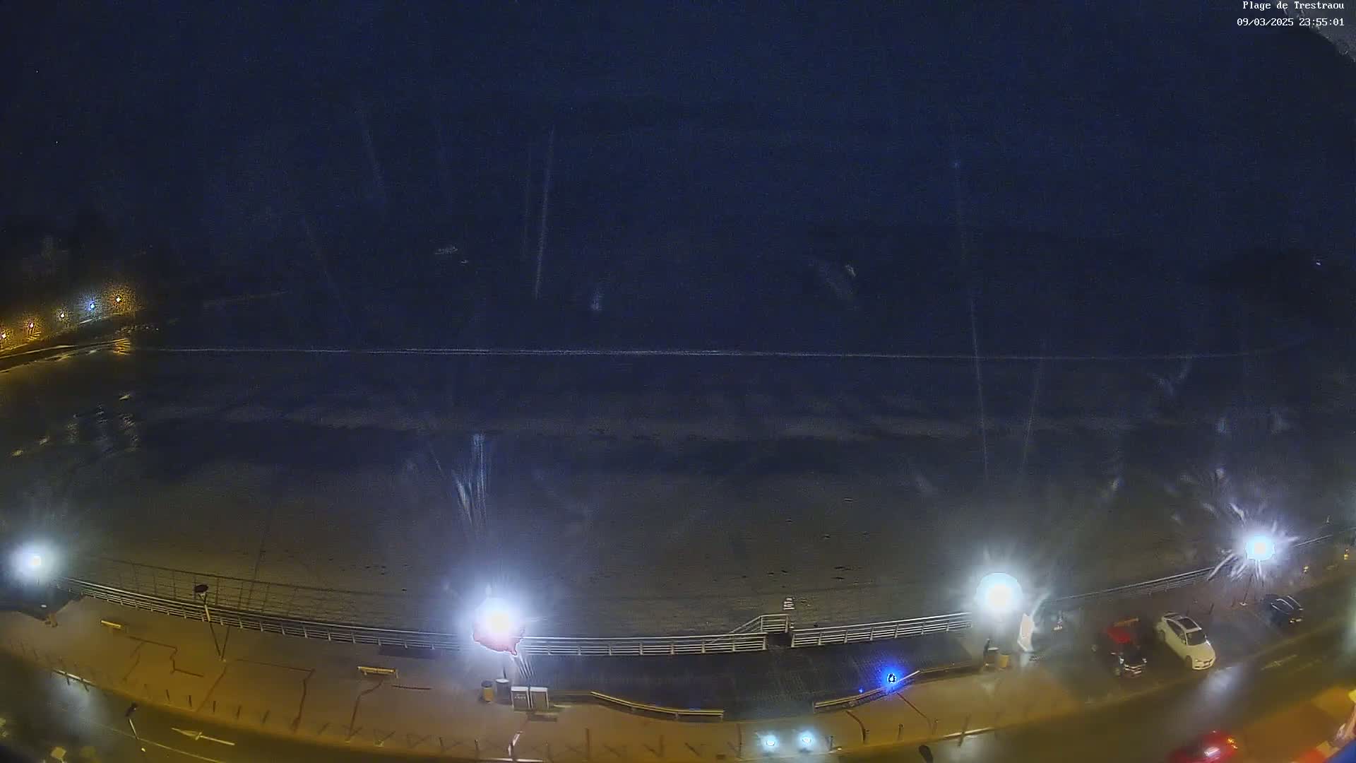 A nighttime, rain-slicked beach and boardwalk are illuminated by streetlights, with a few cars parked along the road.
