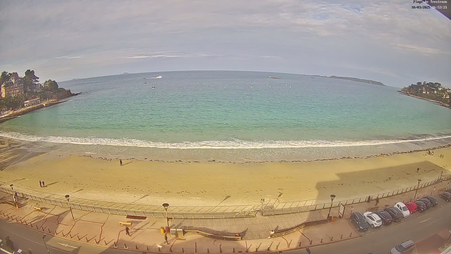 A sandy beach with gentle waves laps at the shore under a partly cloudy sky, with a town and calm ocean visible in the background.