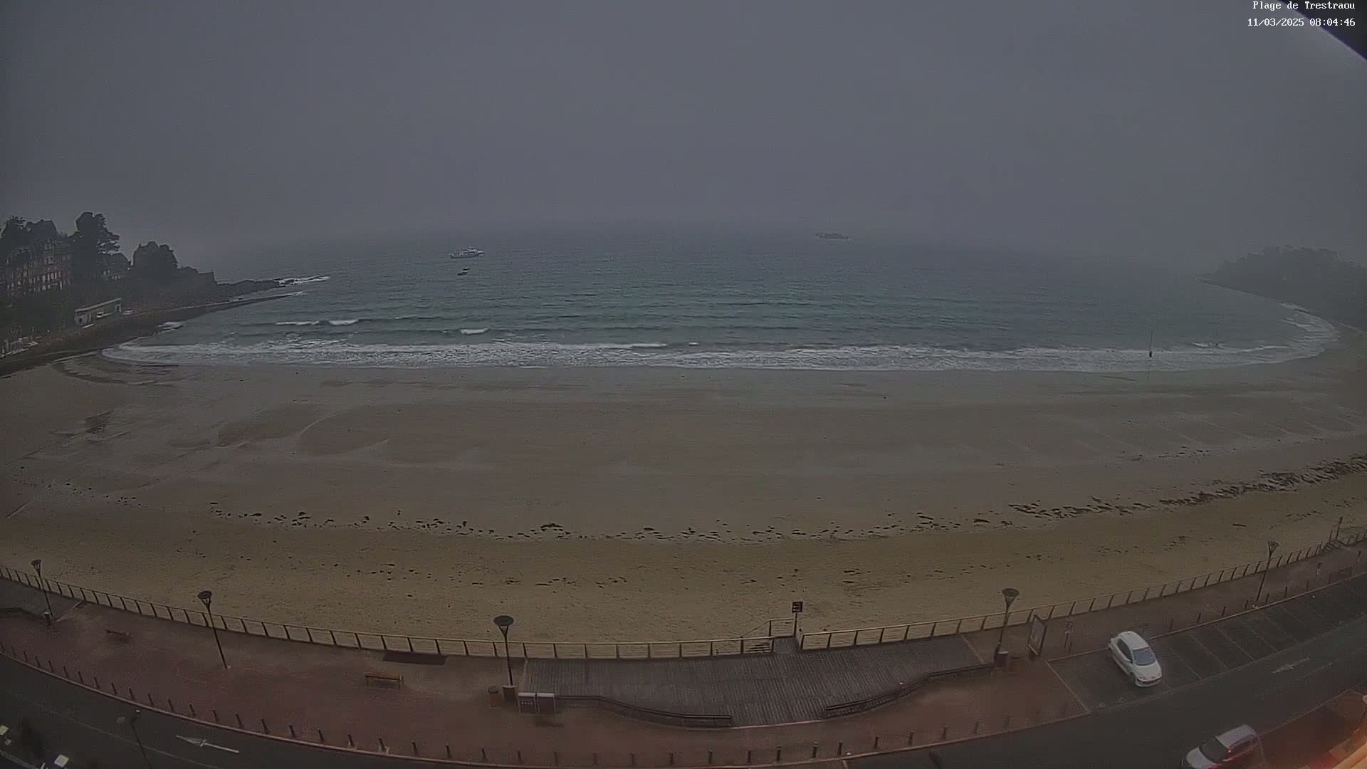 A foggy day reveals an empty sandy beach with gentle waves lapping the shore, overlooked by a paved walkway and a few parked cars.