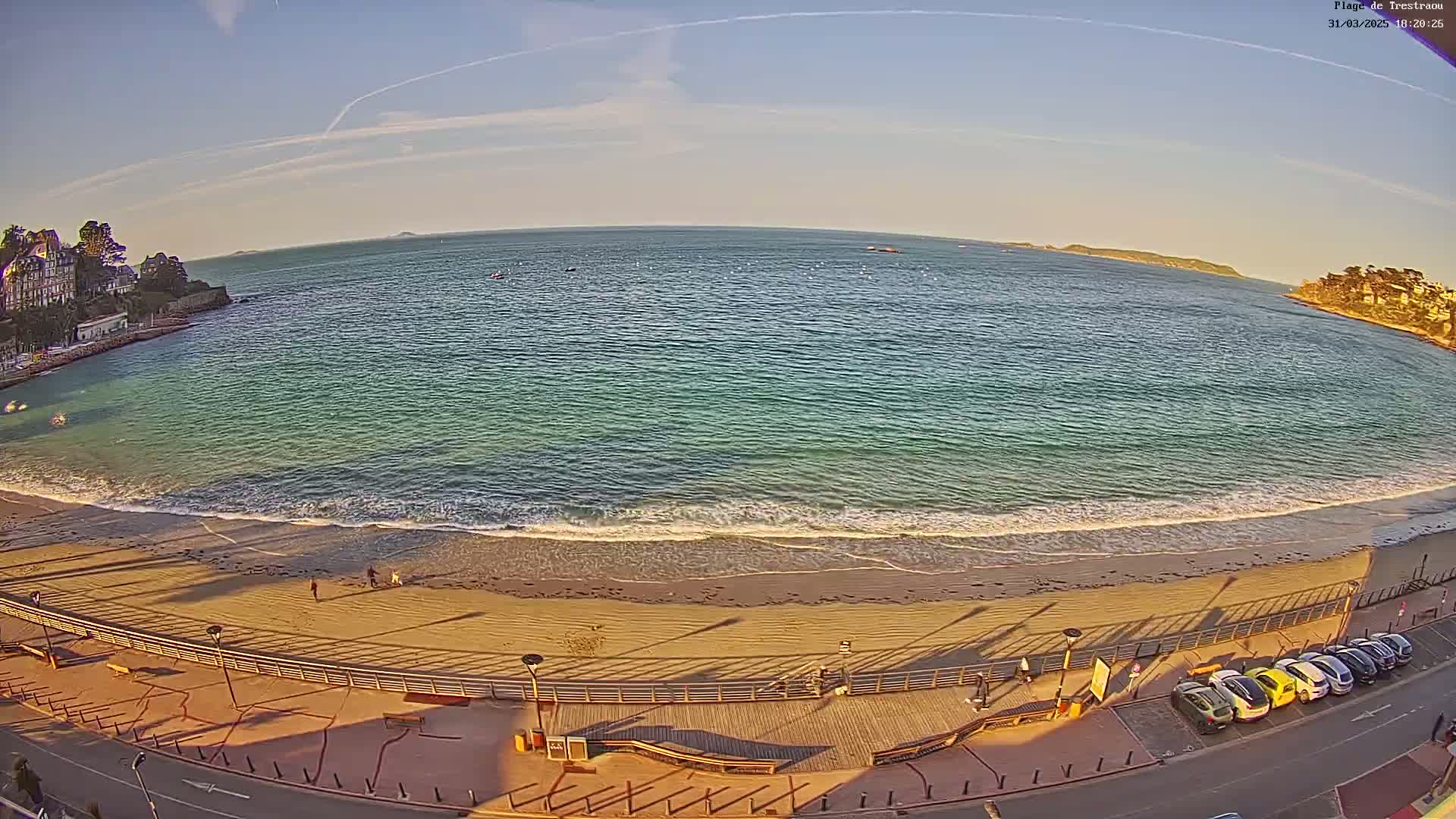 A sandy beach with gentle waves laps the shore under a mostly clear sky, with buildings and parked cars visible along the waterfront.