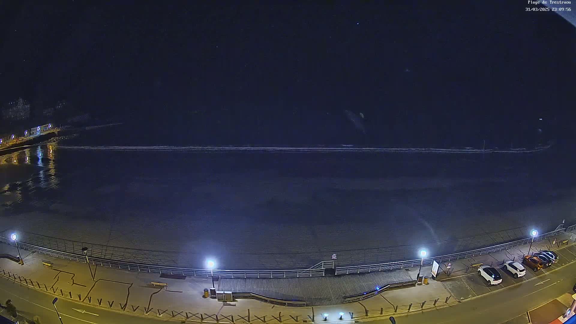 A nighttime, high-angle view shows a dark beach with calm waves under a clear, starry sky, next to a boardwalk with parked cars along a roadway.