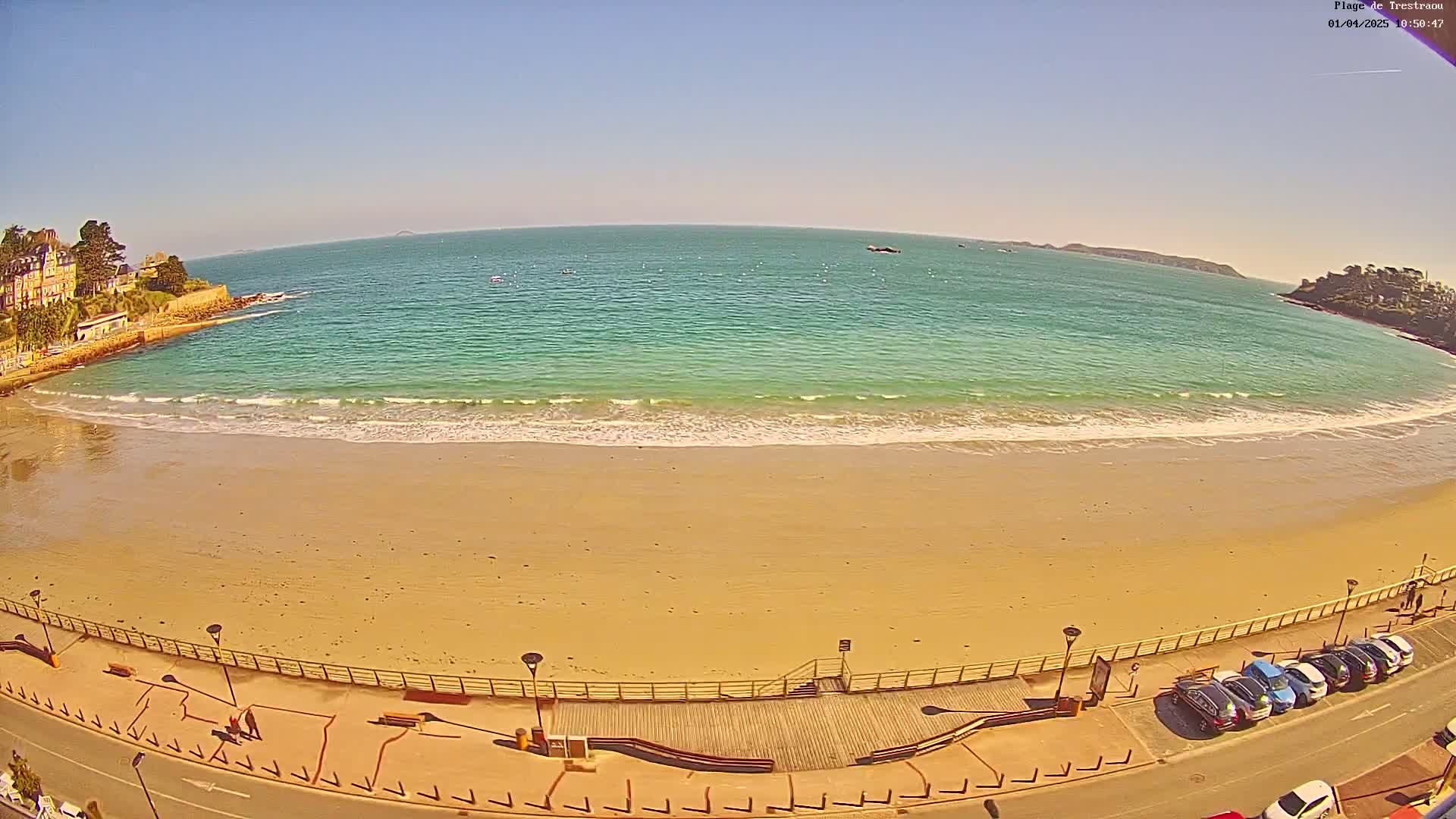 A sandy beach with gentle waves laps the shore under a clear blue sky, with buildings and parked cars visible along the beachfront.