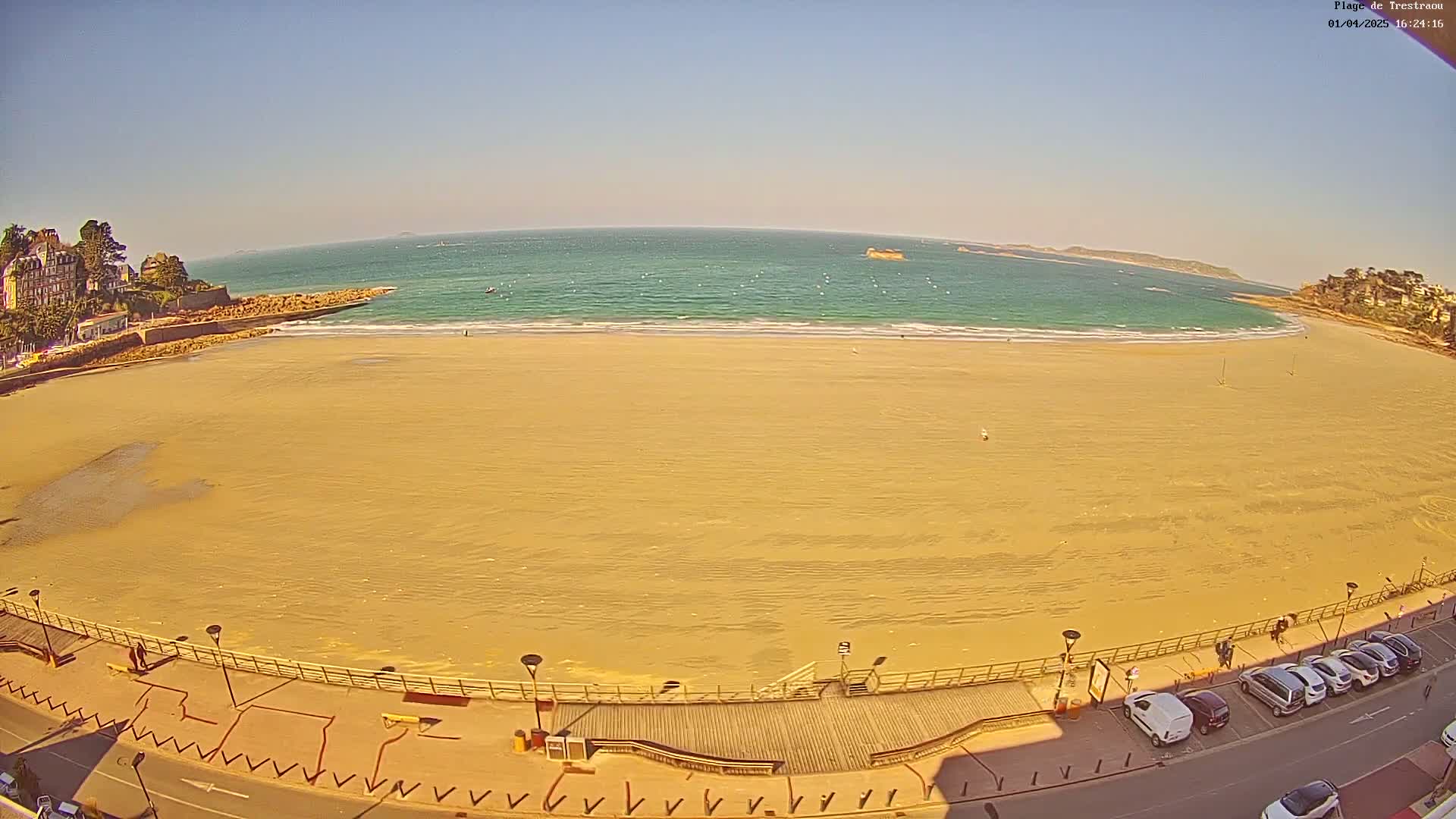 A wide sandy beach stretches along a calm, teal ocean under a clear blue sky, with buildings and parked cars visible along the shore.