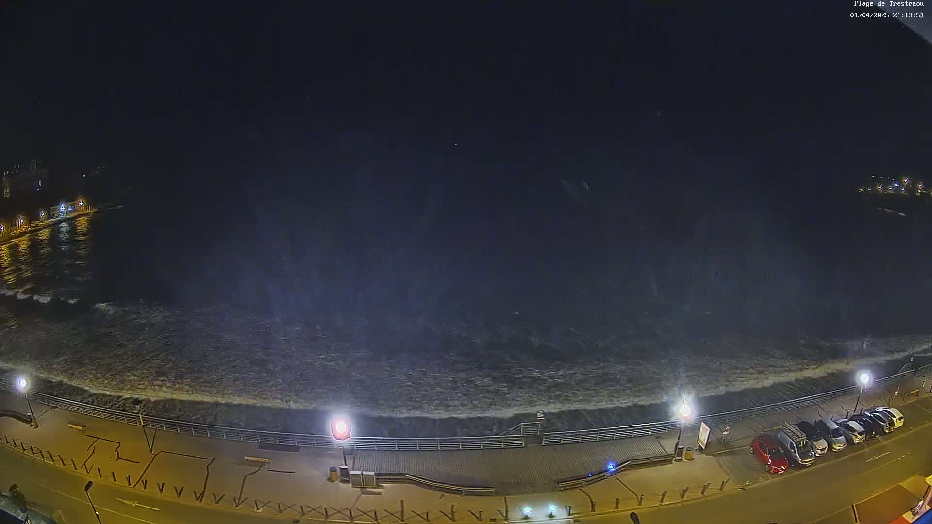 A nighttime, high-angle view shows a dark ocean with moderately sized waves crashing against a boardwalk with parked cars and streetlights along a paved road.