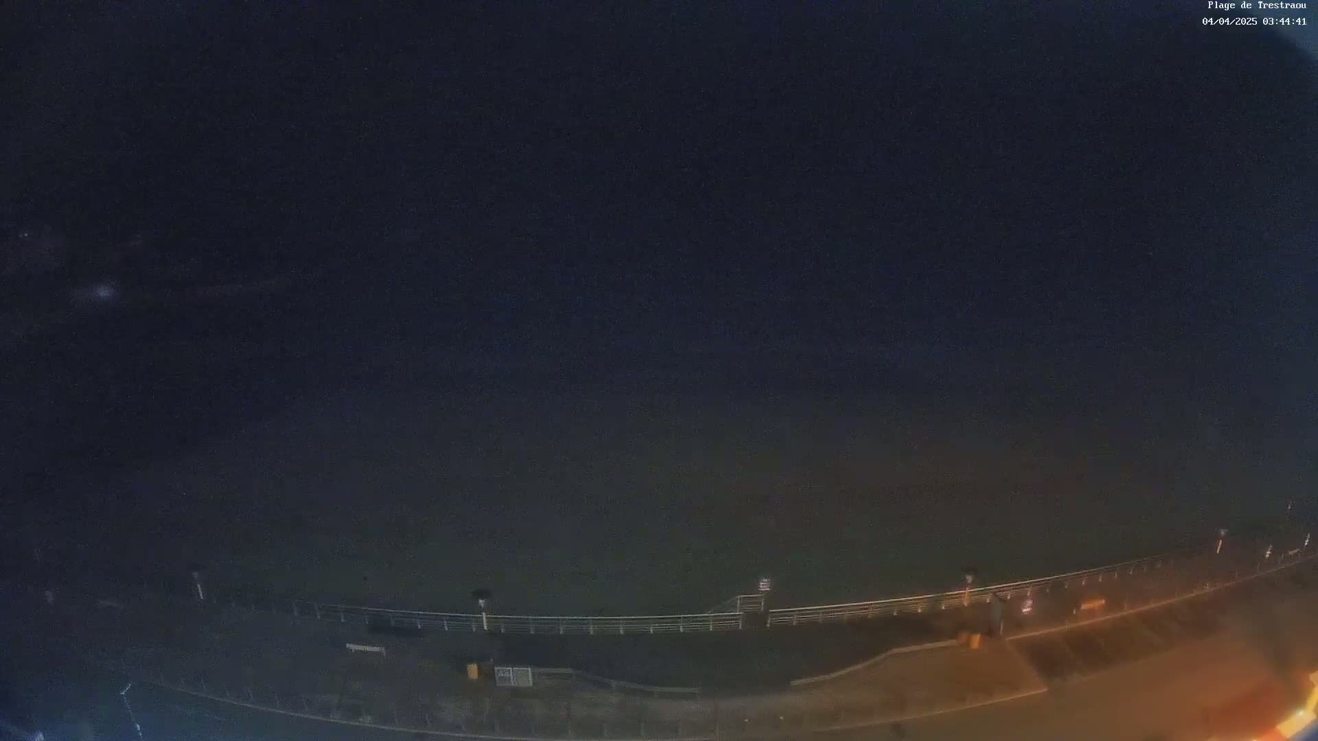 A nighttime, high-angle view shows a dark body of water next to a paved area with a railing, a few lights, and some people.