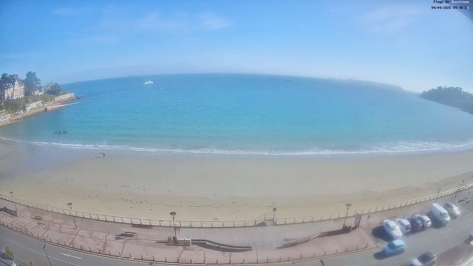 A sandy beach with calm, light-blue water stretches along a coastline under a clear blue sky, with a few cars parked along the road beside it.