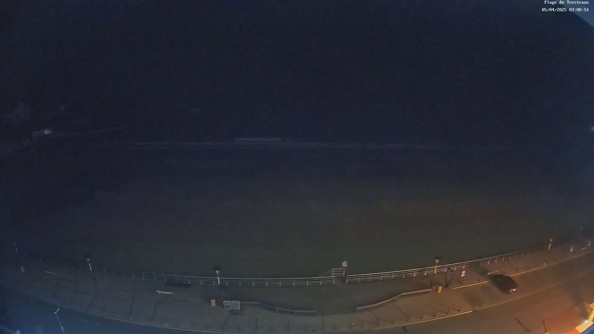 A nighttime, high-angle view shows a dark body of water meeting a paved waterfront with a railing, streetlights, and a single parked car.