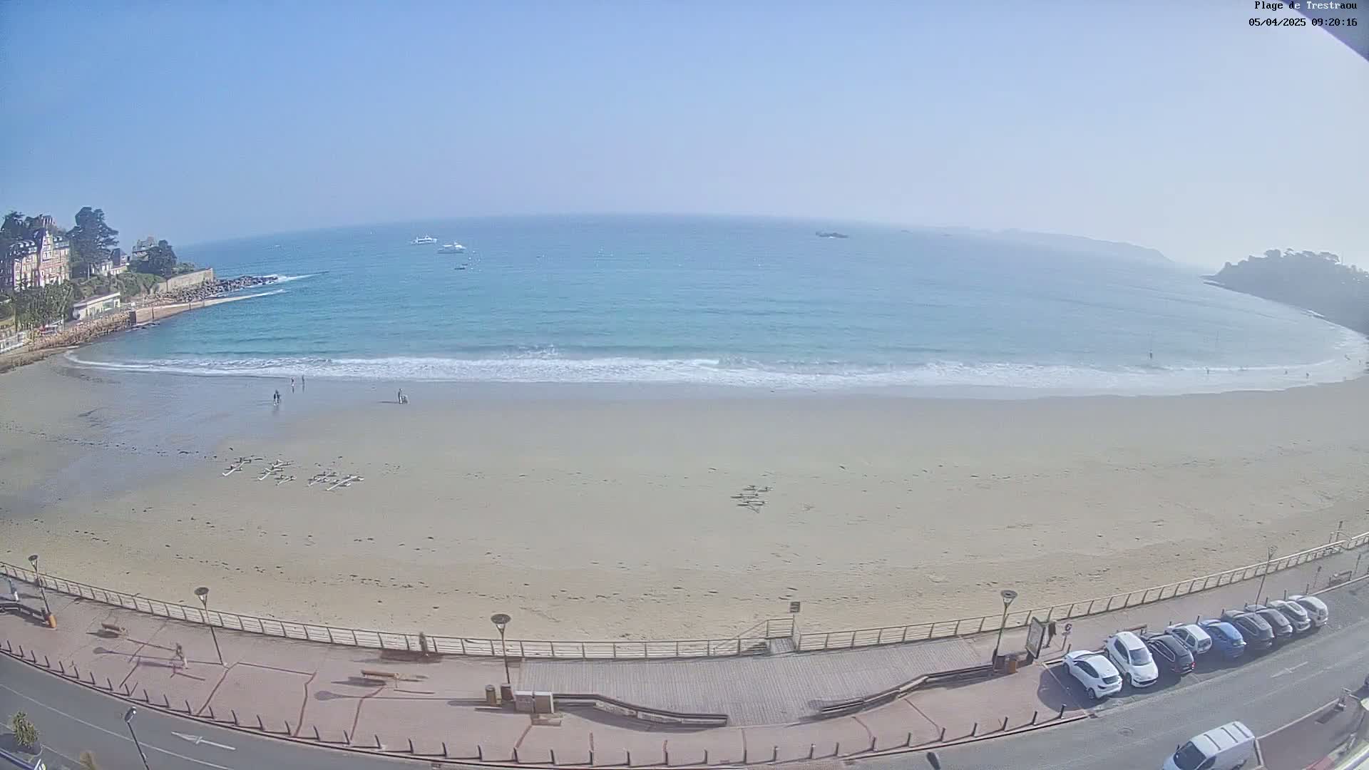 A sandy beach with calm waves laps the shore under a clear blue sky, a few people are visible on the beach and several cars are parked along the road behind a boardwalk.
